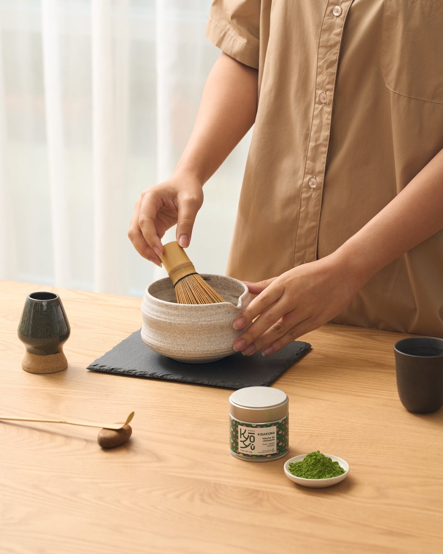 Deux mains élégantes d'une femme, la main droite tenant un fouet chasen en bambou, tandis que l'autre repose délicatement sur le côté d'un bol chawan traditionnel. Un support à chasen est visible sur la gauche.
Notre matcha Kisakuna riche ne Umami et aux notes de noisette est mis en avant avec une coupelle remplie de matcha en poudre