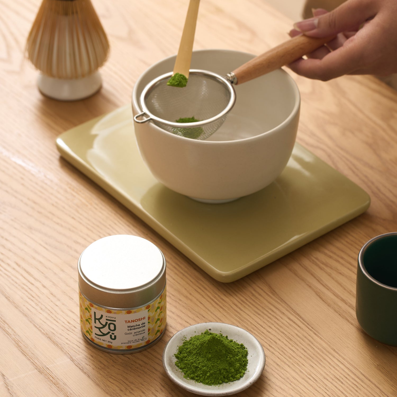 Matcha Tanoshi posé sur une table avec une coupelle remplie de poudre de matcha, tandis que les mains d'une femme préparent un matcha avec soin.