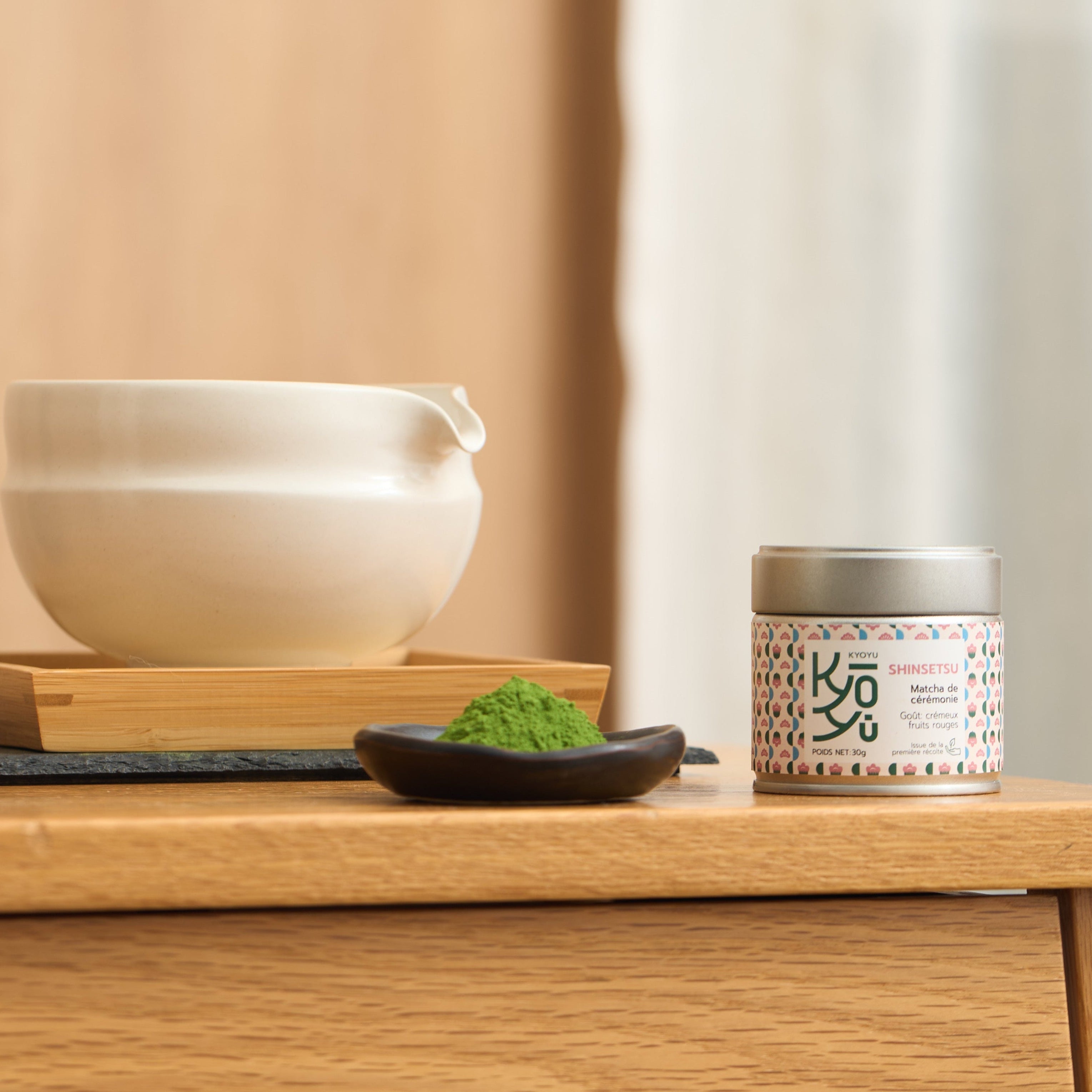 Notre matcha Shinsetsu, posé sur une table avec a ses coté une coupelle remplie de poudre de matcha. En arriere plan, un bol a matcha de couleur blanche. 