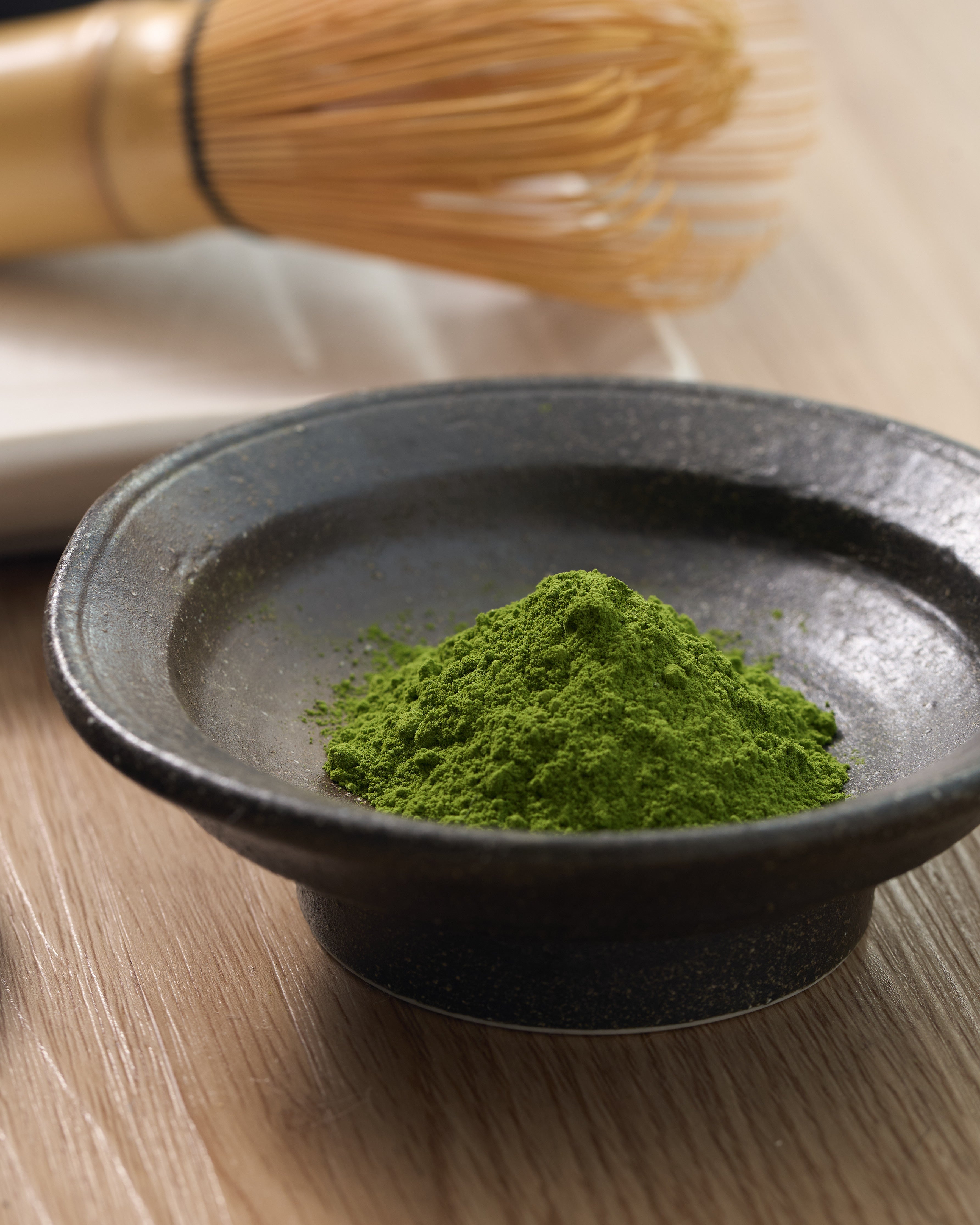 photo zoomée dune coupelle en céramique remplie de matcha en poudre d'une couleur verte éclatante. En arrière plan et un peu floutée, on distingue un fouet chasen traditionnel.