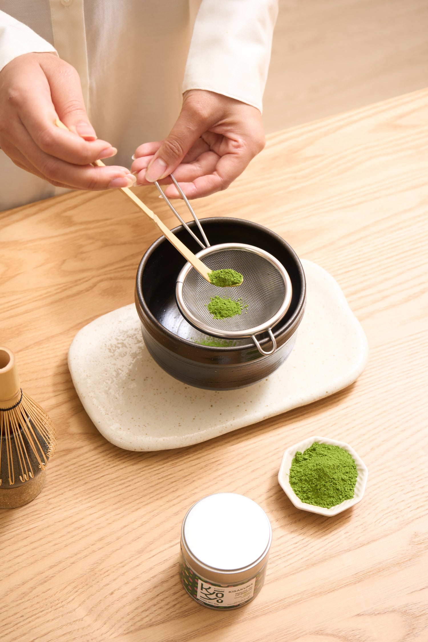 du matcha dans une cuillere en bamboo en train d'Être tamisé au dessus d'un bol hawan. Un fouet en bamboo se trouve à côté.