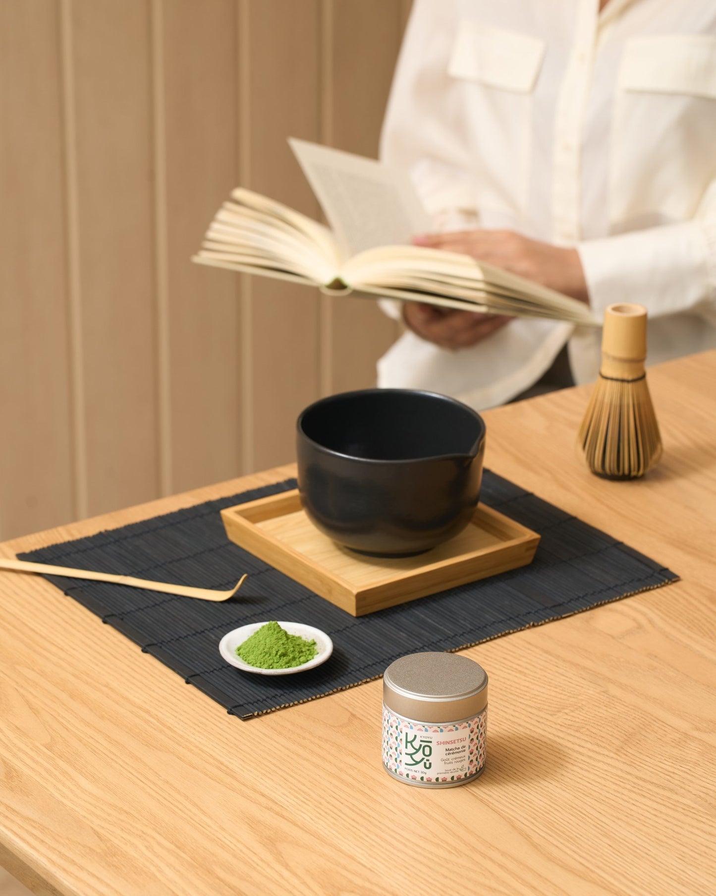 Le matcha Shinsetsu est mis en avant. En arrière plan on retrouve un chawan et un chasen posés sur la table. Une femme est assise et tient un livre dans ses mains. 